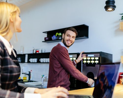 Shot of a young couple talking in an office cafeteria. Man making a coffee.